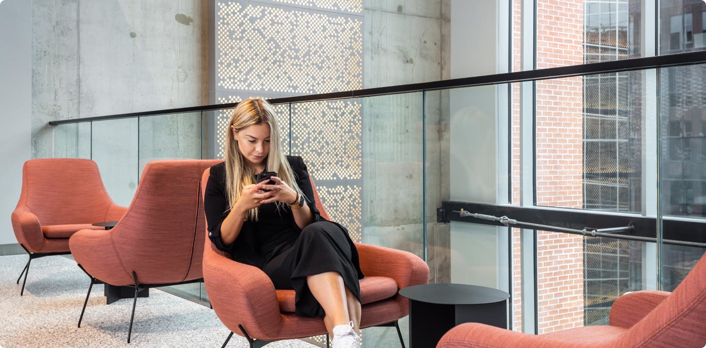 Woman sitting on a pink chair in on a mezzanine in a Citybox Hotel, looking at her phone.